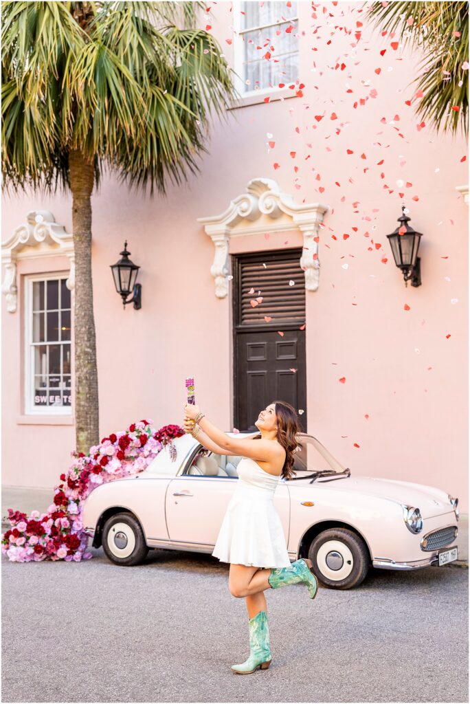 A Charleston senior in a white dress with green cowgirl boots, popping red and white confetti during her senior photos with The Pink Figgy, who is dressed in a floral garland from The Charleston Petal, at the Mills House Hotel on Meeting Street in Charleston, South Carolina