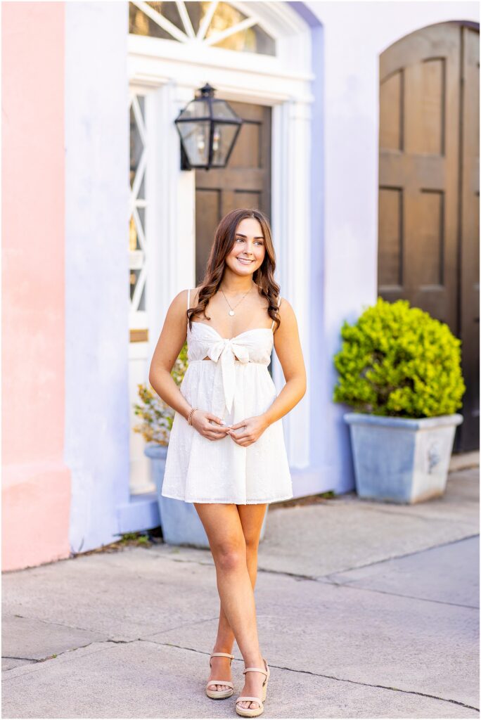 Pastel homes creating a beautiful backdrop for a high school senior posing in a white dress on Rainbow Row in Charleston