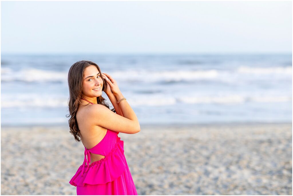 Senior girl posing on Folly Beach with her hands in her hair as she wears a bright pink dress