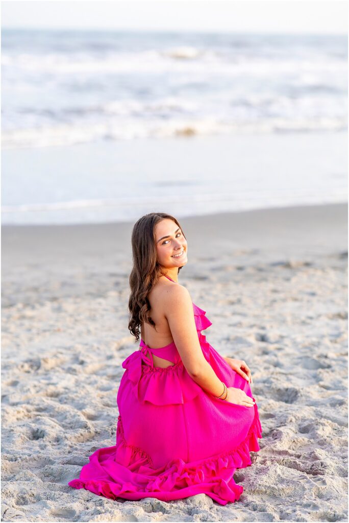High school senior girl crouching down in the sand on Folly Beach, wearing a bright pink dress and looking over her shoulder as she poses for photos
