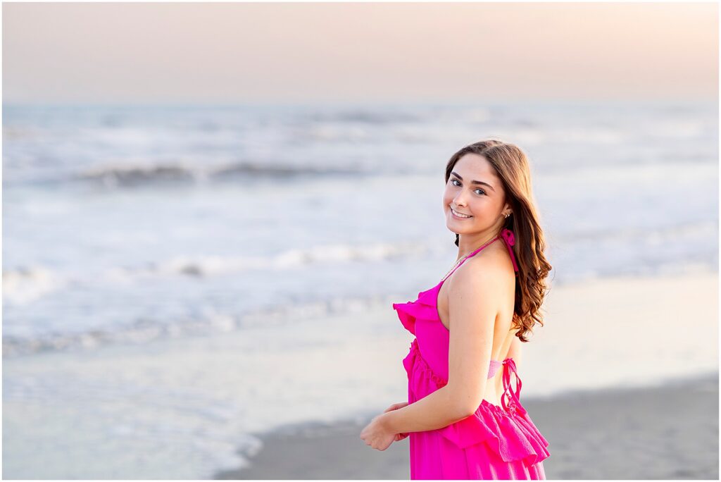Timeless senior portrait of a senior girl in a hot pink dress with the ocean sunset as the backdrop on Folly Beach, South Carolina