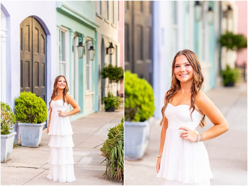 Charleston senior smiling in front of pastel homes with her hand on her hip while wearing a white dress for her senior photos