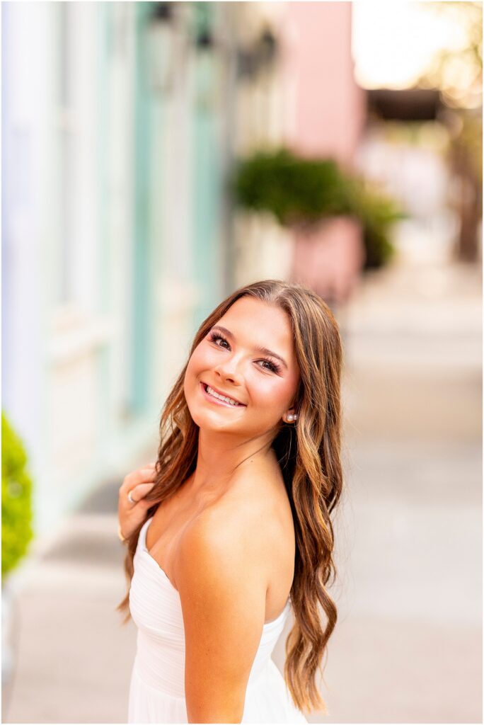Charleston senior girl smiling for a portrait on Rainbow Row with pastel homes blurred behind her
