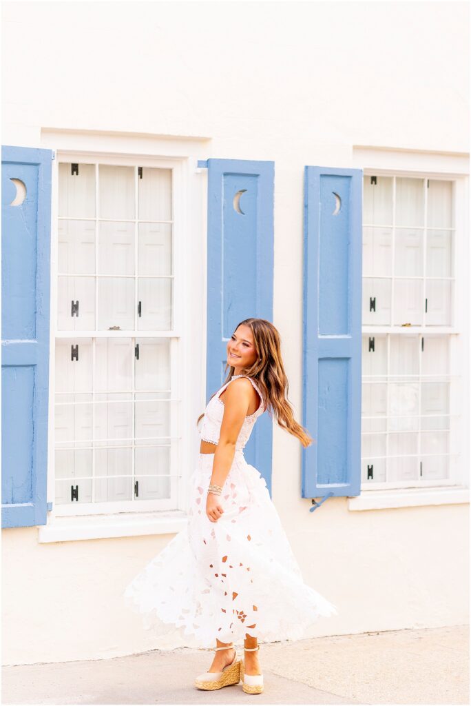 Fun image of a senior girl twirling in her dress in front of a white home on Tradd Street in Charleston with blue shutters