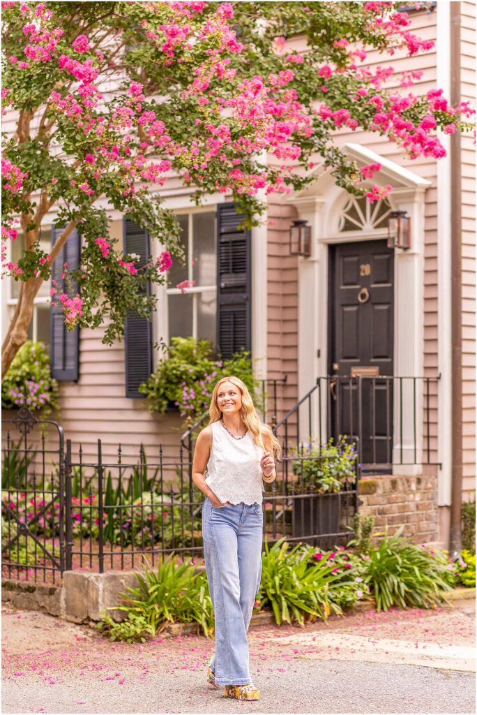 High school senior in a white top and jeans walking through the South of Broad neighborhood in Charleston with blooming pink flowers