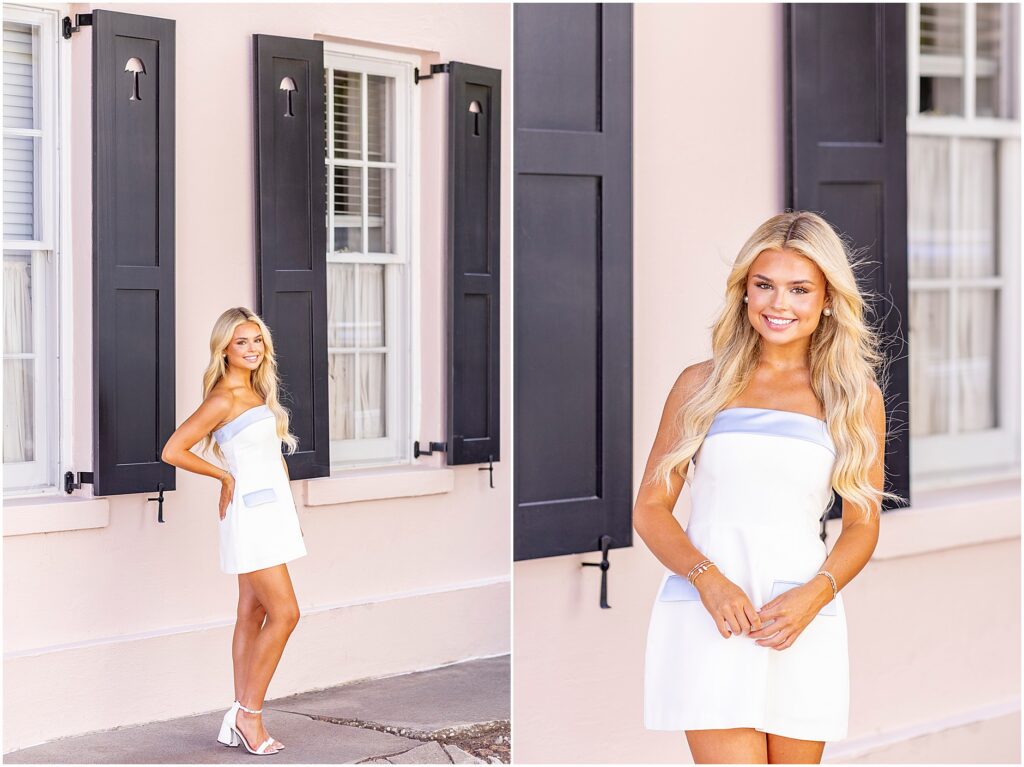 Senior girl in a short white dress with blue trim posing in front of a pink Rainbow Row home in Charleston, South Carolina