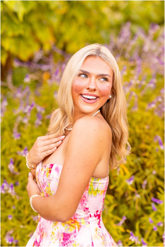 Senior girl in a bright pink dress posing among colorful purple flowers and lush greenery at Hampton Park in Charleston SC